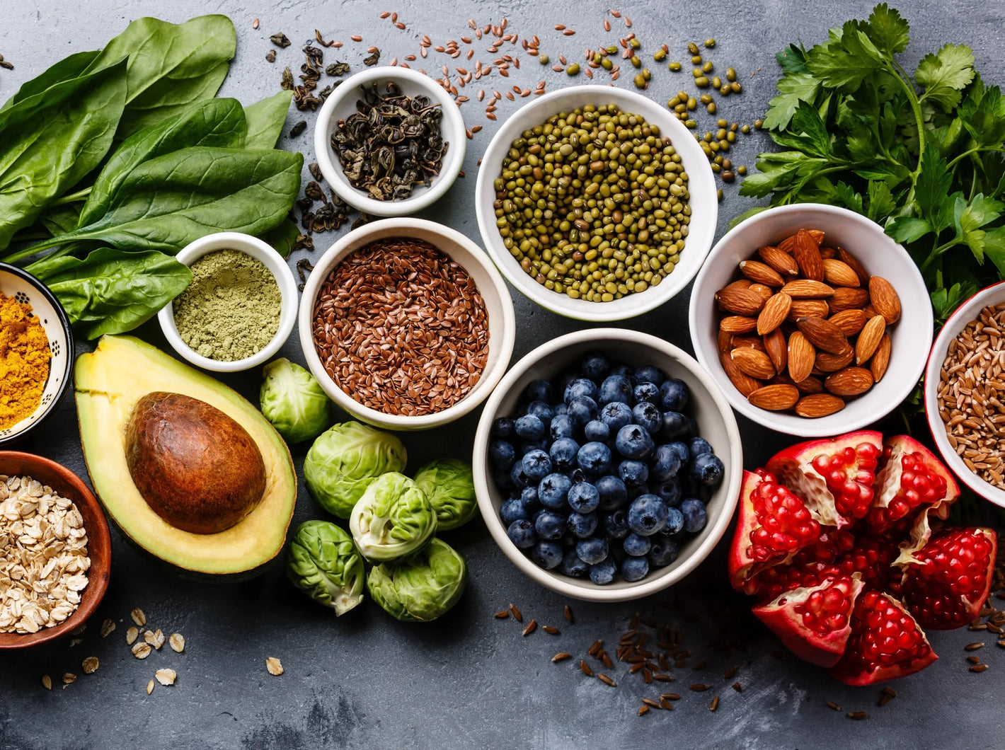 Assorted superfoods including spinach, avocado, blueberries, nuts, seeds, grains, and pomegranate on a gray surface.